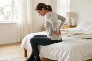 Femme assise sur le bord d'un lit, mains dans le creux des reins, illustration du lumbago