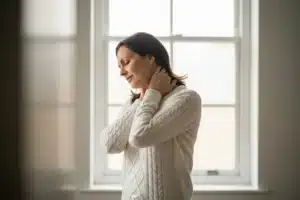 Femme se tenant le cou, soulagée après un torticolis, debout devant une fenêtre lumineuse
